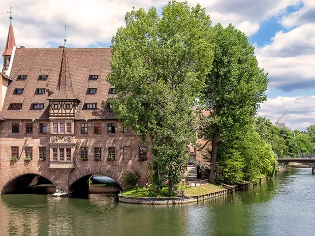 Regionales Ortsmotiv: historisches Gebäude mit Rundbögen und rotem Dach am Wasser neben einem großen Baum.