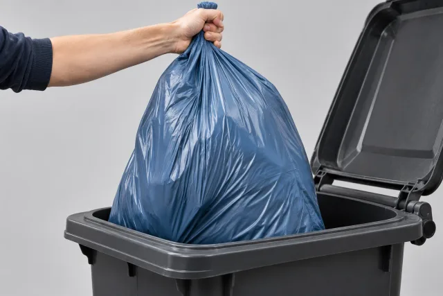 Eine Hand hebt einen gefüllten blaugrauen Müllsack am gerafften Hals aus einem dunklen Abfallbehälter.